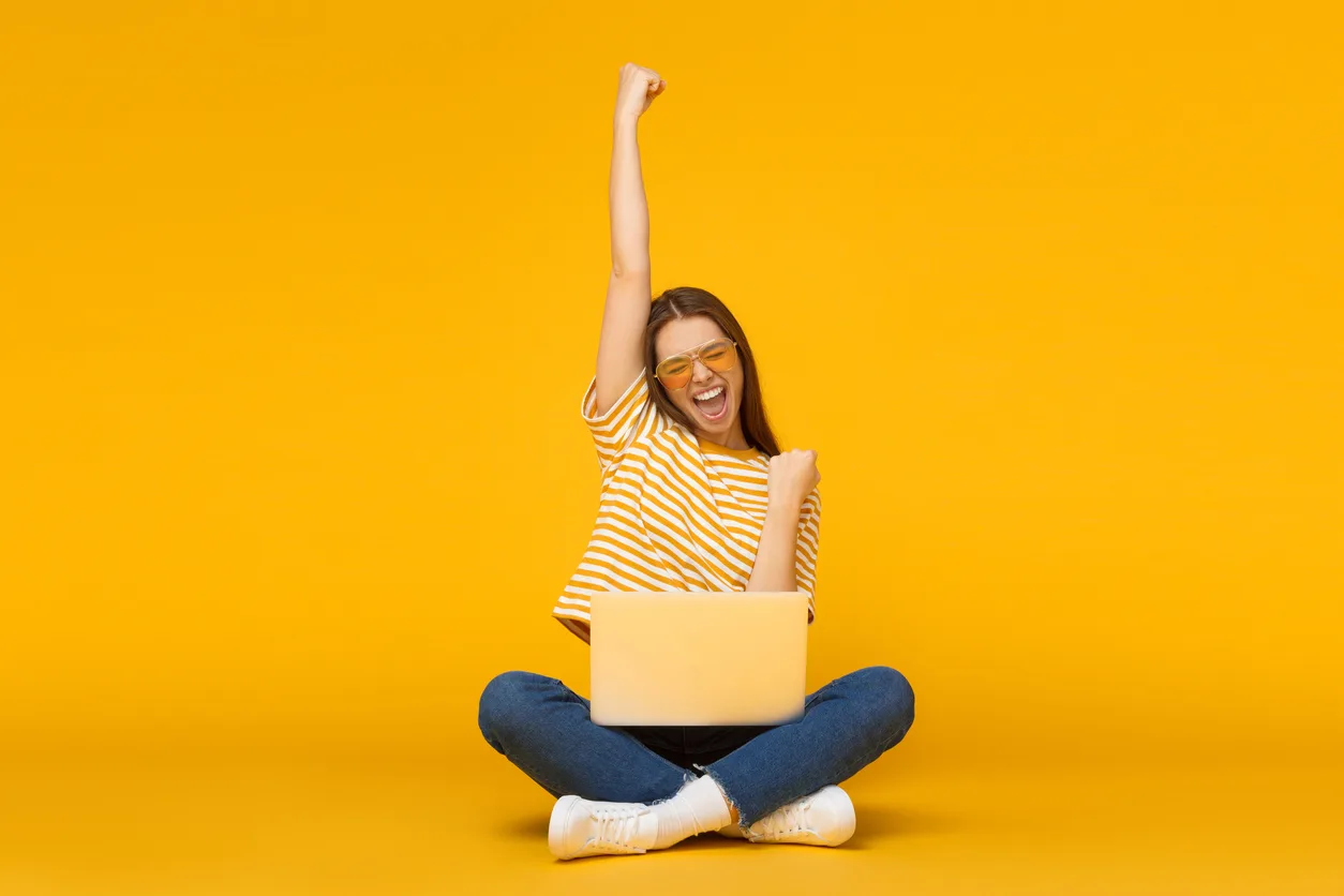 A young woman sitting cross legged with a laptop cheers with her arm raised on a yellow background.